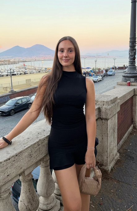 A young woman in a black sleeveless dress posing by a stone balustrade overlooking a harbor and city skyline at dusk, with Mount Vesuvius visible in the background.
