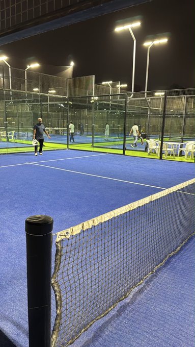 An evening view of an outdoor padel or tennis court complex with blue playing surfaces, chain-link fencing, and bright floodlights, where several people are visible playing or standing on the courts.