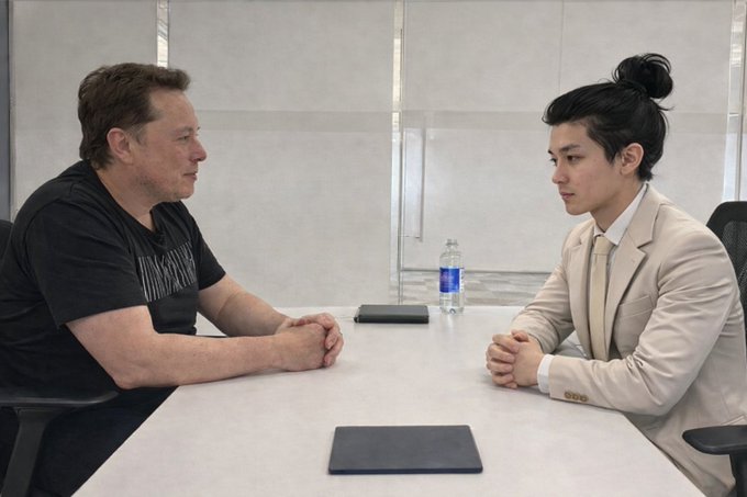 Elon Musk, dressed casually, has a meeting at a conference table with a younger man in a beige suit whose hair is styled in a top knot.