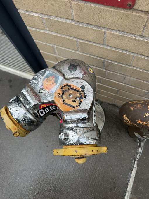Two weathered silver fire hydrants covered in stickers (including 'OBEY' and a pixelated character) sit on a sidewalk against a tan brick wall.