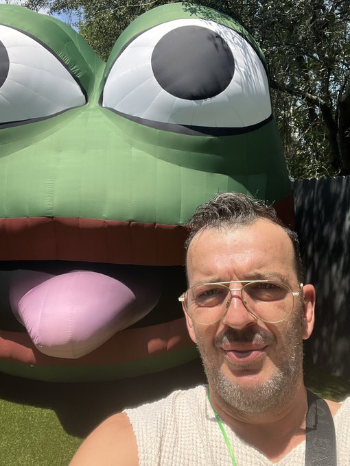 A selfie of a man with glasses smiling in front of a large inflatable green frog character with prominent eyes and a pink tongue.