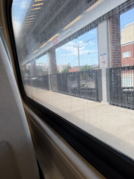 A view from inside a moving train, looking through a speckled window at a station platform and buildings outside.