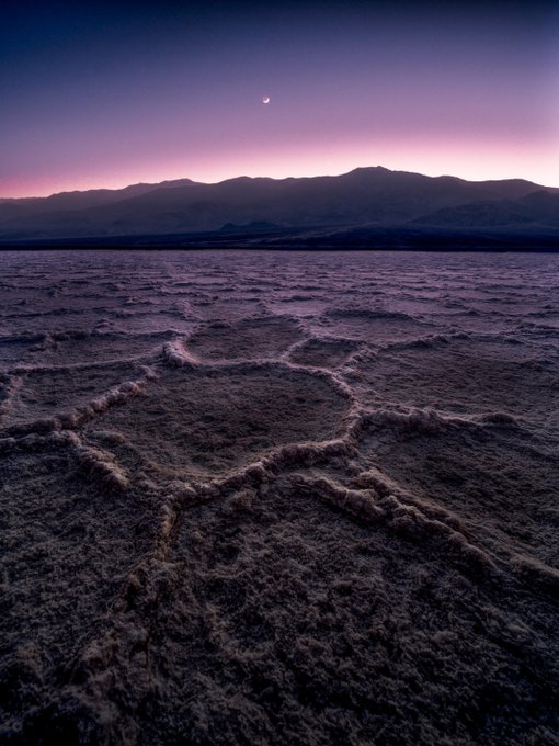 A twilight landscape photograph of the salt flats featuring a stick-figure geoglyph in the foreground, with mountains silhouetted against a purple and pink sky and a small crescent moon above.