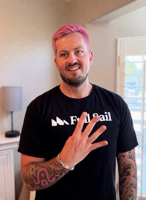 A man with pink hair and a beard smiles at the camera while making a hand gesture, wearing a black 'Full Sail' t-shirt with visible arm tattoos.