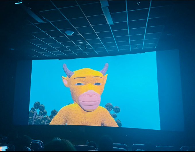 A photograph taken inside a movie theater showing a large screen displaying a 3D animated yellow cow character with horns and a pink snout.