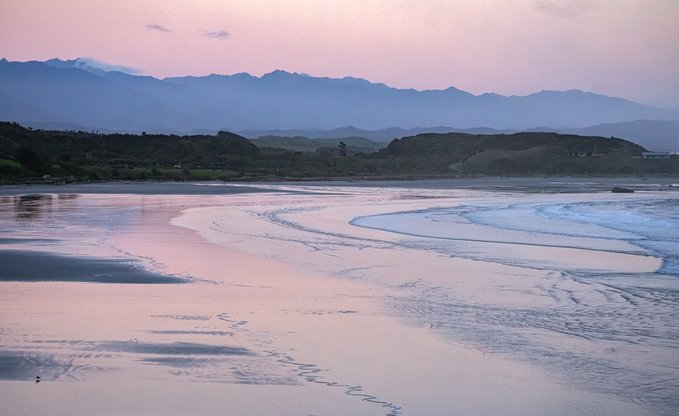 A serene coastal landscape at dusk with wet sand reflecting pink and purple hues, gentle waves rolling onto the shore, and mountains visible in the background.