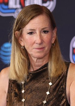 A woman with blonde hair wearing a brown velvet dress and a long beaded necklace, photographed at what appears to be an NBA All-Star event.