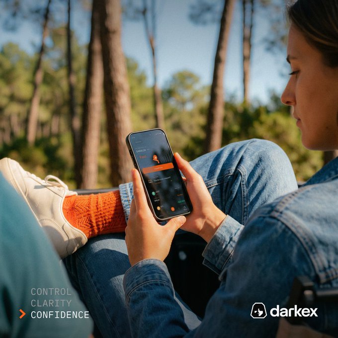 A person sits outdoors in a forest in casual clothing in orange in a focus mode in a focused in a confident in a control and a clarity and a confidence in a control on a darkex app.