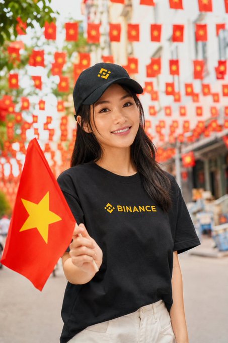 A smiling woman wearing a black "BINANCE" t-shirt and matching cap holds a Vietnamese flag, standing on a street decorated with strings of red and yellow flags.