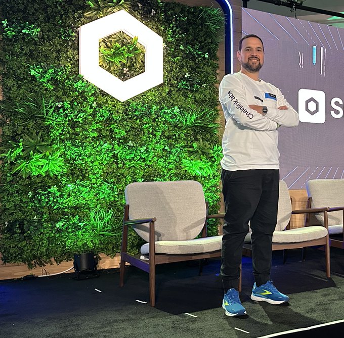 A man in a white "Chainlink Labs" shirt poses confidently with arms crossed in front of a green living wall featuring a hexagonal logo and a screen displaying the Chainlink branding, visually celebrating his new role managing Chainlink Labs' Community Advocate Program.