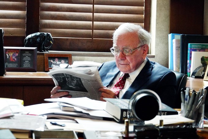 An elderly gentleman in a suit and red tie sits at a cluttered office desk, intently reading a newspaper, with a "Berkshire" nameplate visible among the paperwork and books. The image accompanies a post questioning Warren Buffett's unexpected sale of $285 billion in Japanese bonds, framing his careful study of financial news as potentially revealing insider insight before Japan's debt figures were released.