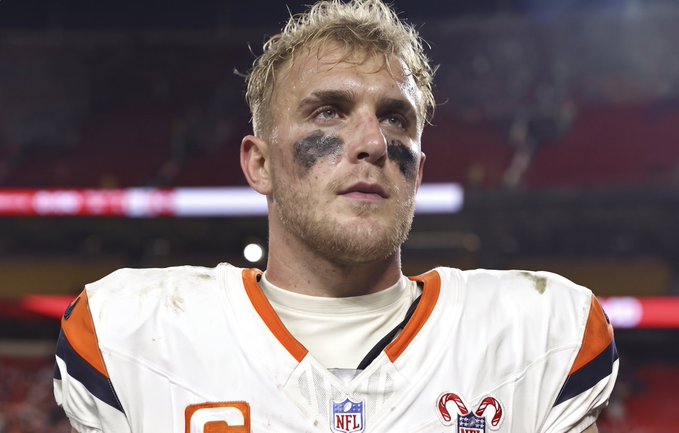 A young blonde football player with black eye black stands on the field wearing a white Denver Broncos jersey featuring the NFL shield and a captain's "C" patch, captured in a contemplative moment that aligns with the caption "Bo Nix in 2026" suggesting a future veteran look for the Broncos quarterback.