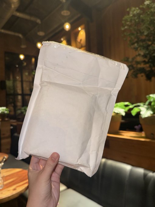 A hand holding up a white sealed pouch/package in what appears to be a cafe or restaurant setting with plants and warm lighting in the background.