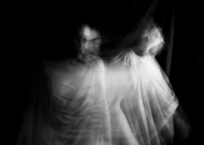 A haunting, ghostly black-and-white image of two figures that appear blurred and ethereal, shrouded in translucent, web-like material against a dark background.