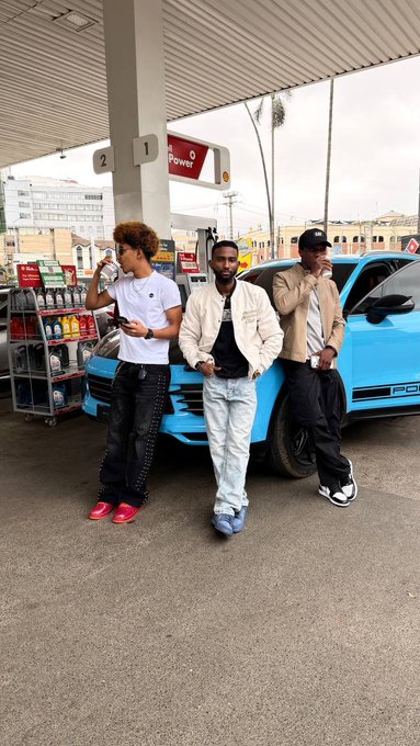 Three young men pose in front of a bright blue Porsche at a gas station with pumps labeled "1" and "2," reinforcing the post's call for followers to join the "XM boys and girls" trading community as they prepare for Monday's live YouTube session in Kenya featuring a prominent local trader.