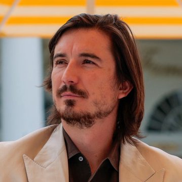 A man with shoulder-length brown hair and a goatee, wearing a tan blazer over a brown shirt, is shown in a thoughtful pose, though no text or identifiers are visible in the image to connect it to Robinhood or the $HOOD tokenized assets context.