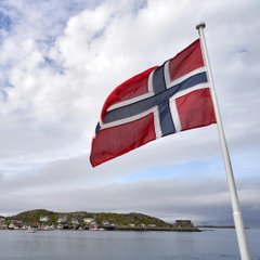 A Norwegian flag waves in the wind against a coastal backdrop of islands and sea, symbolizing Norway as the source of the news that the country's sovereign wealth fund has reached an all-time high indirect Bitcoin exposure of 11,549 BTC (~$725M) in H1 2026.