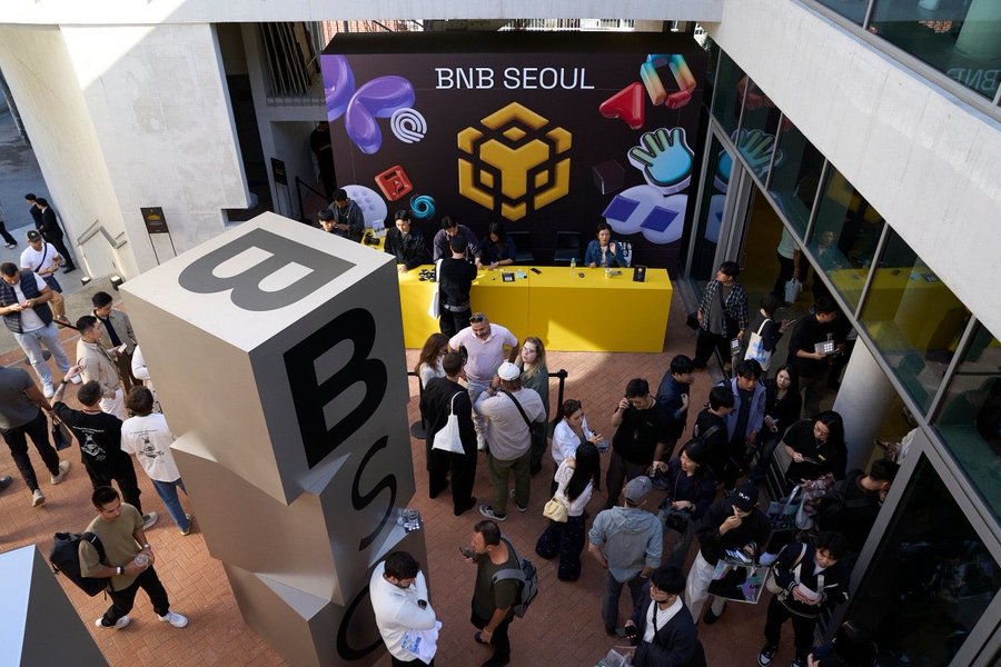An overhead view of a crowded BNB Seoul event, featuring a giant BNB cube sculpture and attendees gathered around a yellow booth.