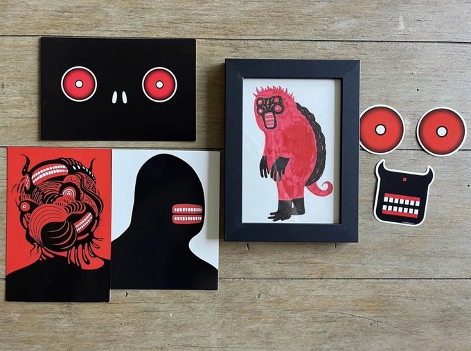 A collection of artistic prints, stickers, and cards on a wooden surface featuring surreal black and red monster-like figures with prominent teeth and eyes, including a framed print of a red furry creature.