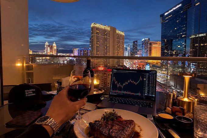 A person holds a glass of red wine at a balcony dinner overlooking the Las Vegas Strip at dusk, with a laptop displaying a stock chart on the table and a black cap reading 'Discipline Equals Freedom' nearby.