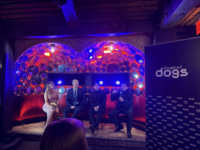 A photo of a panel discussion at a 'dogs' event (appears to be the kaspa/doginals community), featuring four people seated on stage with colorful lighting and a 'dogs' backdrop.