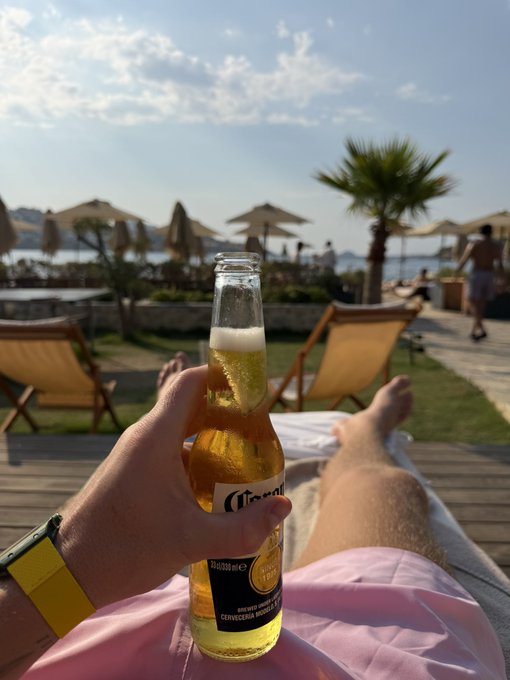 A hand holds up a cold Corona beer bottle (visible label reading "Corona" and "Cerveza") in a toast to the day, while the photographer relaxes barefoot on a lounge chair at a beachside resort with palm trees and umbrellas in the background. The casual scene perfectly captures the carefree "Good morning I am alive" spirit — embracing a leisurely, sun-soaked start to the day.