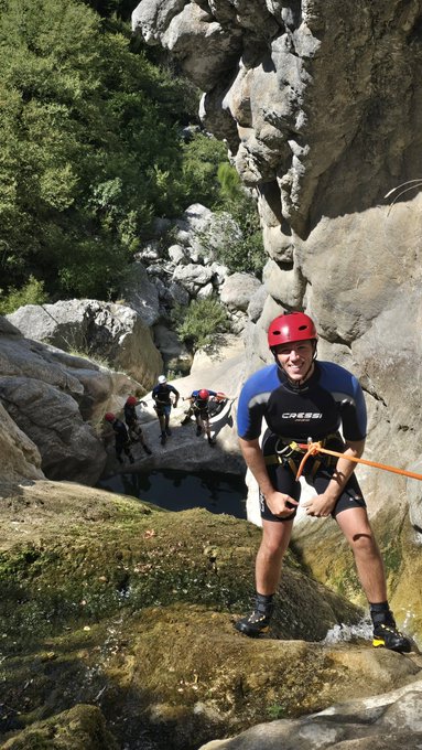 A smiling man in a red helmet, sunglasses, and a 'Cressi' wetsuit stands on wet rocks in a canyon, holding an orange rope with other people visible in the background.
