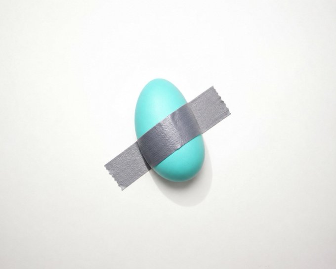 A minimalist photograph showing a light blue egg-shaped object wrapped with a single strip of gray duct tape, resting on a plain white surface.