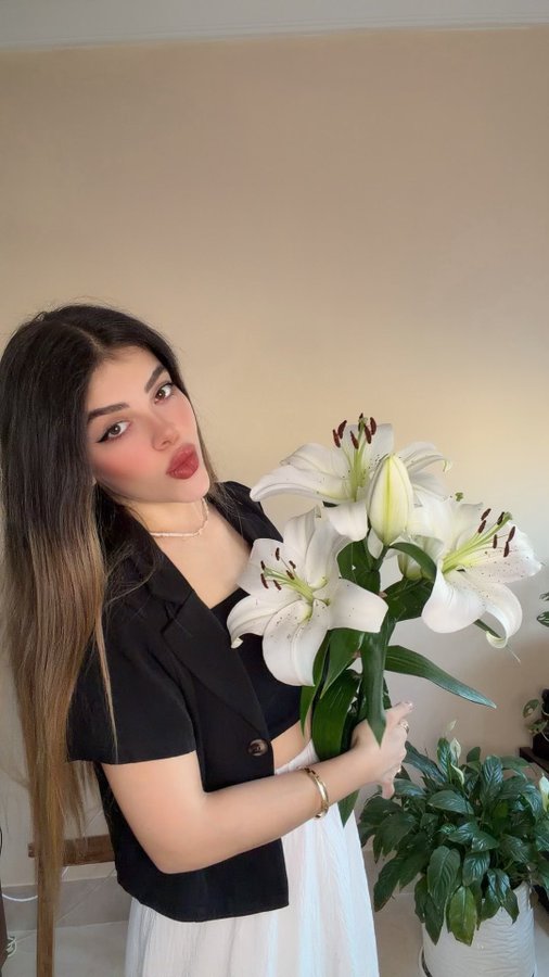 A young woman wearing a black blazer and white skirt holds a bouquet of large white lilies and poses for a portrait indoors.