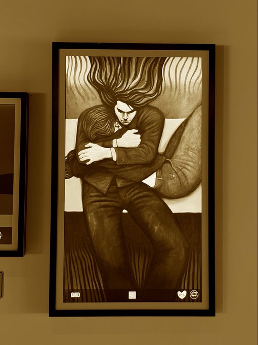 A framed sepia-toned artwork depicting a figure with wild flowing hair embracing a smaller form, displayed on a wall with a partial view of another framed picture beside it.