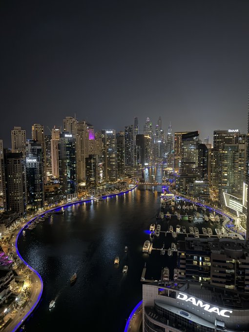 A nighttime aerial view of a Dubai marina with illuminated skyscrapers, boats docked in the harbor, and the DAMAC building prominently visible in the foreground.