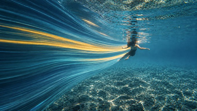 A silhouetted figure floats near the ocean surface while flowing streaks of golden and blue light sweep through the water, capturing the artwork's theme of "light becoming motion" and water serving as a living canvas as described in the Rising #593 drop.