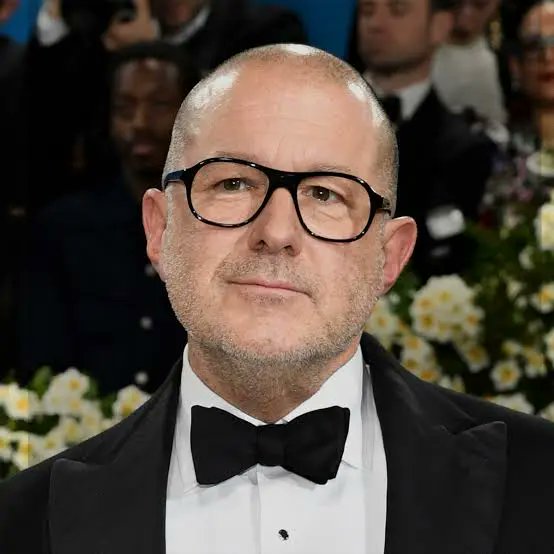 A man in a black tuxedo with a bow tie and glasses poses at a formal event with photographers in the background. The post humorously references Jony Ive (Apple's iconic designer) while seeking a design visionary to create a sleek, premium neobanking platform in crypto.