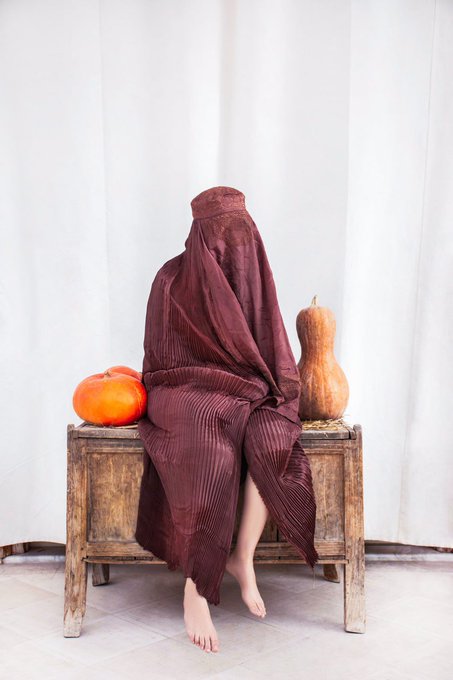A person draped in a flowing, deep red/burgundy garment with their head completely covered sits on a rustic wooden bench, flanked by an orange pumpkin on one side and a tan butternut squash on the other, against a white curtain backdrop.