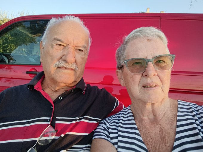 A selfie of an older couple posing together in front of a red van; the man has white hair and a mustache wearing a striped polo, and the woman wears green-tinted glasses and a striped top.