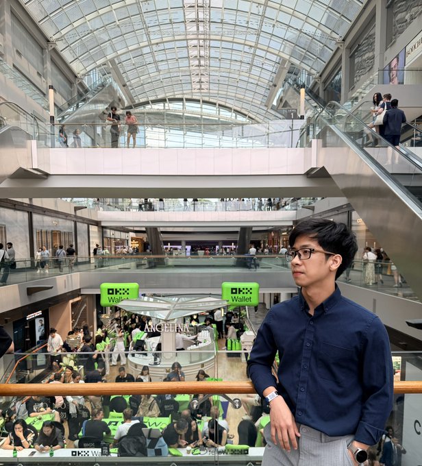 A young man in glasses and a navy blue shirt leans against a railing in a multi-level shopping mall, with digitally inserted green 'PIX Wallet' and 'ANGELINA' signs floating in the scene.