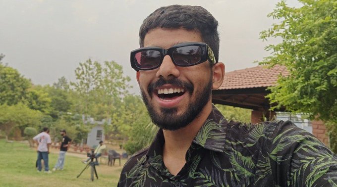 A cheerful man wearing sunglasses and a tropical-print shirt smiles broadly outdoors, with a film crew and tripod visible in the background on the grassy set. The image conveys a "gm gm" (good morning) energy, fitting for a post celebrating a small investment gain from $15 to $20.