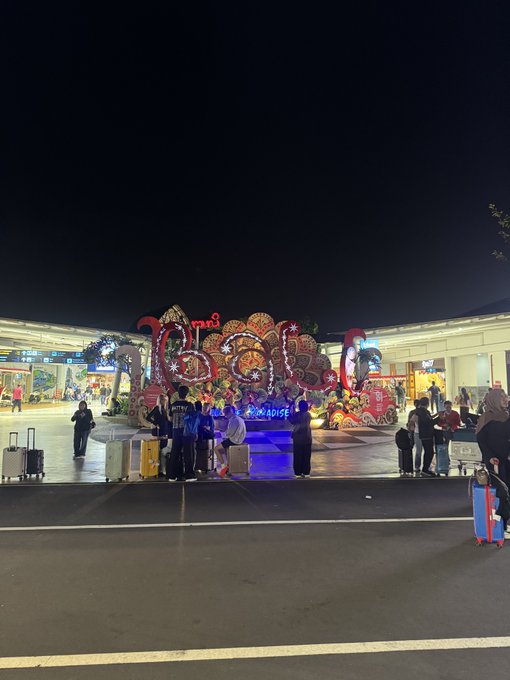 A nighttime photo of a hotel entrance, possibly 'Paradise' resort, decorated with a large illuminated floral display and people with luggage gathered in front.