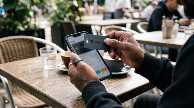 A person sitting at a wooden cafe table holding a Tangerine bank card over a smartphone to make a contactless payment, with a glass of water and a cup of coffee nearby.