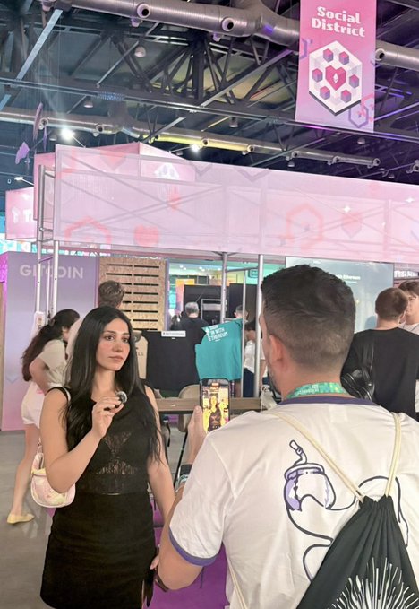 A young woman in a black lace dress holds a microphone while another person records her on a phone at an indoor event with a 'Social District' banner overhead.