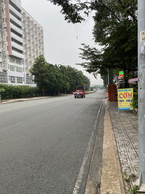 A quiet street scene with a tall white apartment building on the left, trees lining the road, and a red sign advertising 'NHAN COM MAY COM' on the right sidewalk.