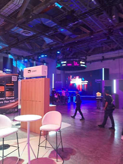 A dimly lit convention hall with blue and purple lighting, featuring a 'Current' booth and people walking around the event space.