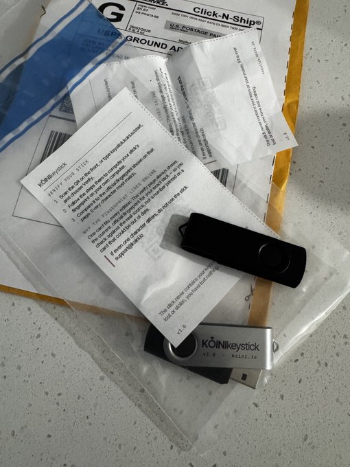 Two USB drives labeled 'KOINtity stick v1.0' lie on top of printed instructions titled 'VERIFY YOUR STICK' and a shipping envelope, appearing to be a package related to a podcast.