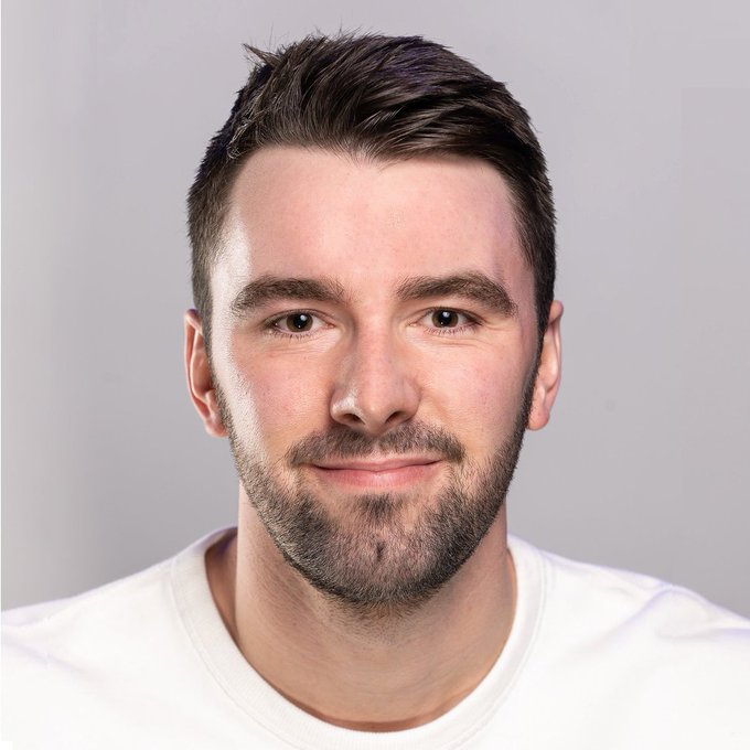 A headshot of a smiling man with short brown hair and a beard, wearing a white t-shirt against a plain gray background.