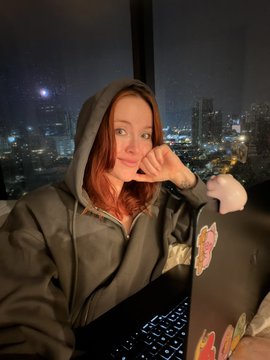 A woman in a dark hoodie sits at a laptop with stickers, making a heart shape with her hand against a backdrop of a nighttime city skyline visible through a window.