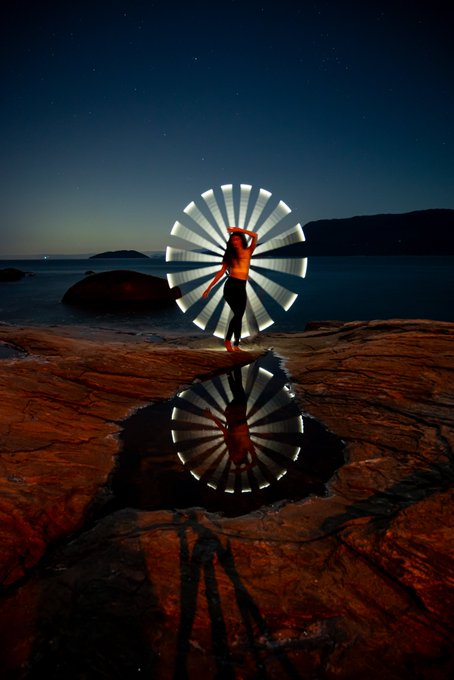 A nighttime long-exposure photograph shows a shirtless person spinning a light source to create a glowing circular pattern, with the illumination perfectly reflected in a tide pool on the rocky shore.