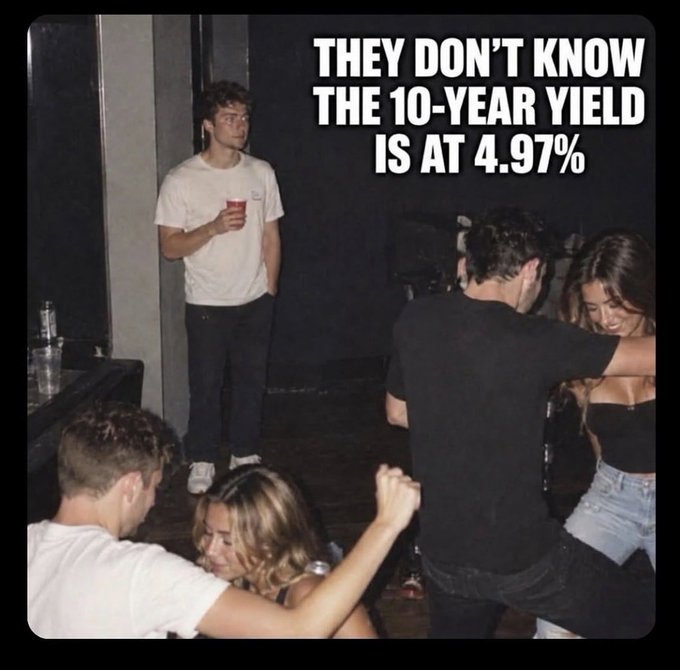 A man stands alone holding a red cup while couples dance around him at a party, with bold text reading "THEY DON'T KNOW THE 10-YEAR YIELD IS AT 4.97%."

The caption humorously captures the "Literally me" sentiment by showing the man's detachment from the social scene—he's not watching the dancers but instead mentally fixated on the notable 10-year Treasury yield figure, portraying a finance nerd oblivious to the party happening around him.