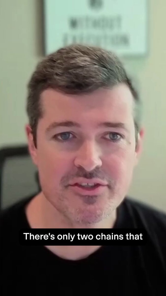 The image features a man wearing a black shirt, with his face clearly visible as he speaks into a microphone. He has a beard and appears to be engaged in conversation or giving a speech. The background of the image is blurred, but there are two chairs present that could potentially be part of an office setting. Additionally, there's a sign on the wall behind the man, which reads "There's only two chains that have been tested at scale..." This suggests that the man might be discussing or talking about a specific product or service related to chains and their testing process.