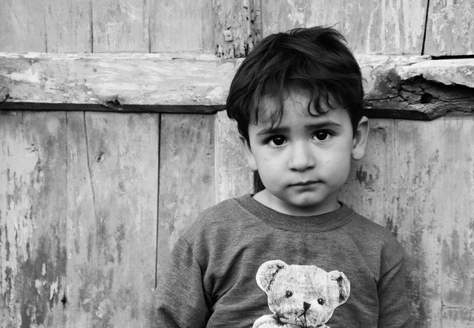 A black and white portrait of a young child wearing a t-shirt featuring a teddy bear graphic, standing in front of a weathered wooden door.
