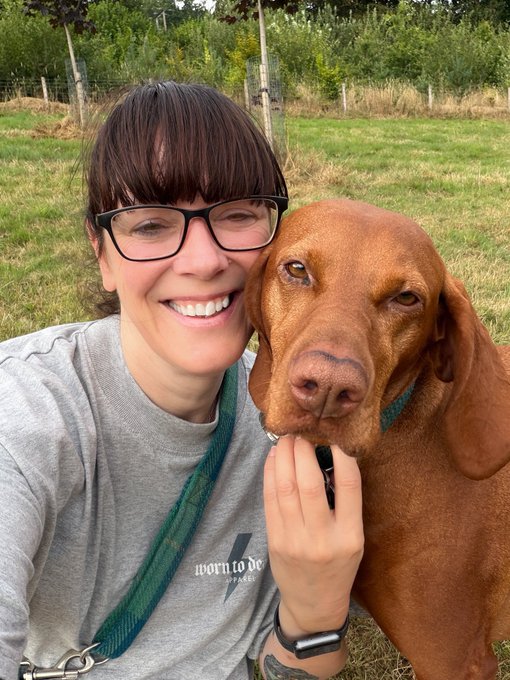 A smiling woman with glasses poses for a selfie outdoors with a brown Vizsla dog resting its head against her face.